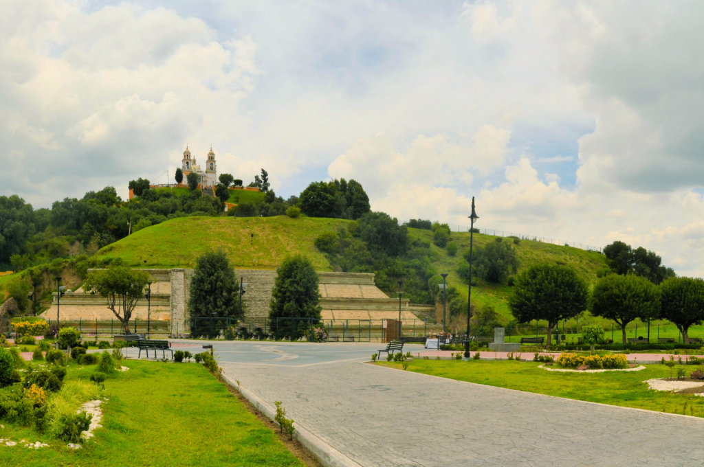 View of the Great Pyramid of Cholula with Nuestra Señora de los Remedios Church on top, surrounded by green landscape.