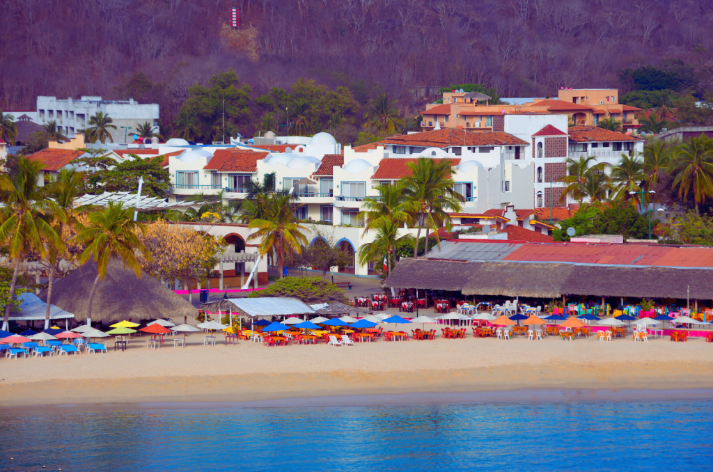 Beachfront view of Huatulco, Mexico, with colorful umbrellas, beachfront restaurants, and resort-style accommodations.