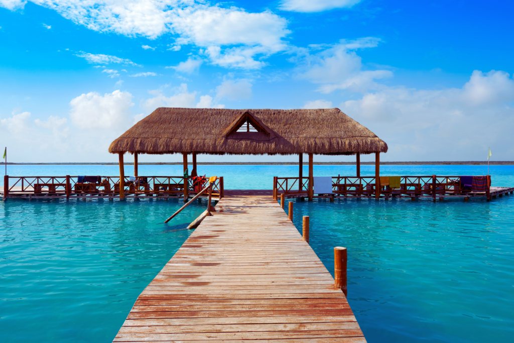 hatched-roof pavilion over the turquoise waters of Lake Bacalar with a wooden walkway leading to it.