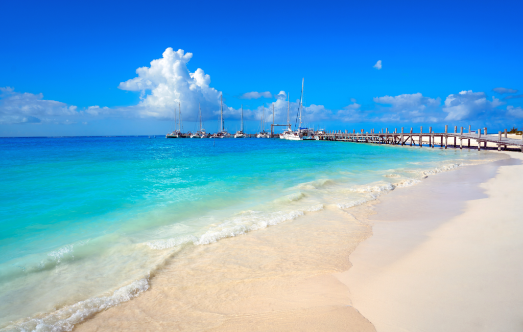 Beach with white sand and turquoise waters with a pier in the background.