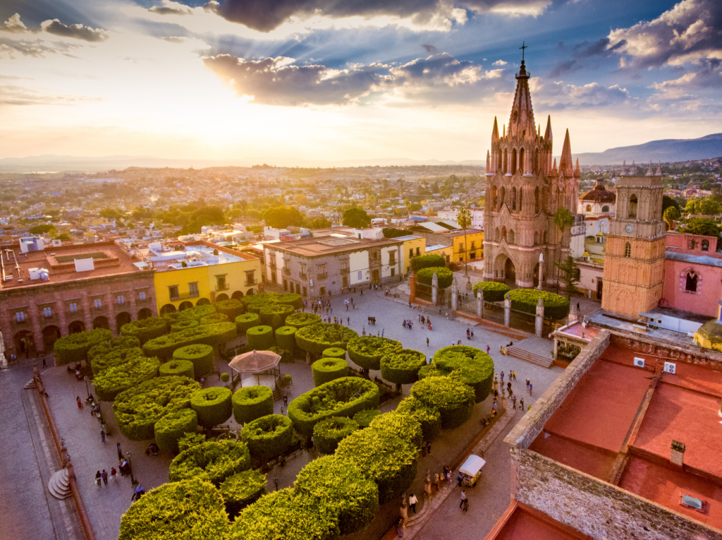 San Miguel de Allende Cathedral and the plaza in front of it, and nearby neighborhoods, with bright sunlight shinning on the rooftops of the houses.