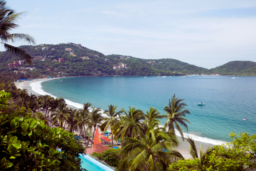 A scenic view of Zihuatanejo's beautiful beach and lush green hills with resort accommodations in the background.